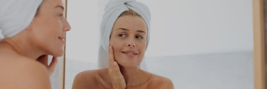 Smiling woman inspecting face in mirror with towel wrapped around head